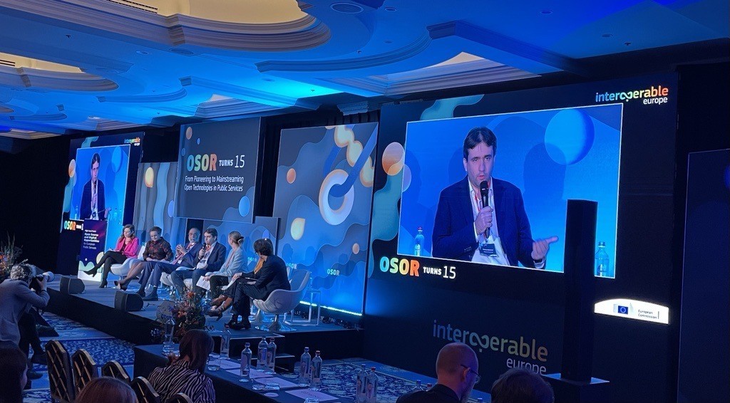 the image shows Bozhidar Bozhanov on two large displays, on either side of the 7 seated panel members