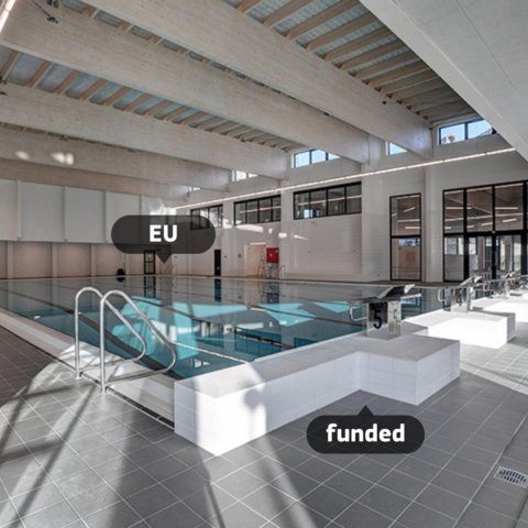 An indoor swimming pool with modern architectural design, featuring large windows and wooden ceiling beams. The pool area is labeled with 'EU' and 'funded' tags, indicating that it was funded by the European Union. 