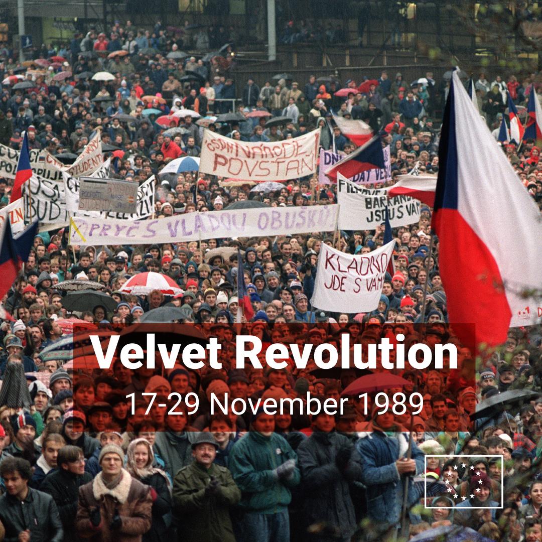 A large crowd of protesters gathered during the Velvet Revolution, many holding banners and Czech flags. People stand closely together in cold weather, some with umbrellas. Overlaid text reads ‘Velvet Revolution – 17–29 November 1989.