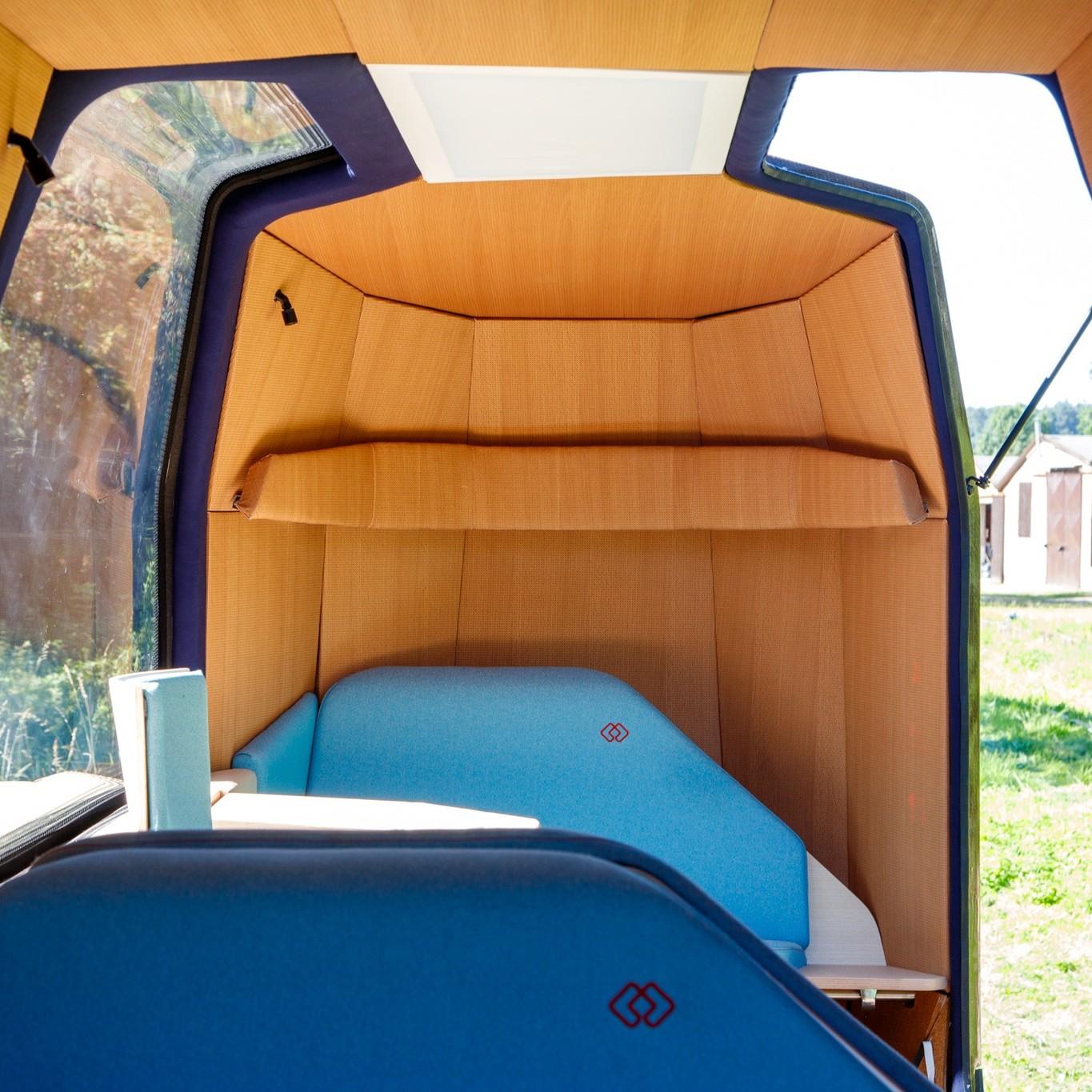 This image shows the interior of a compact, modern vehicle cabin with wooden paneling and large windows. The seating area features blue upholstered seats with a small table or surface nearby. The design is minimalistic and cozy, with natural light coming through the windows, revealing a grassy outdoor scene with a building and trees in the background. 