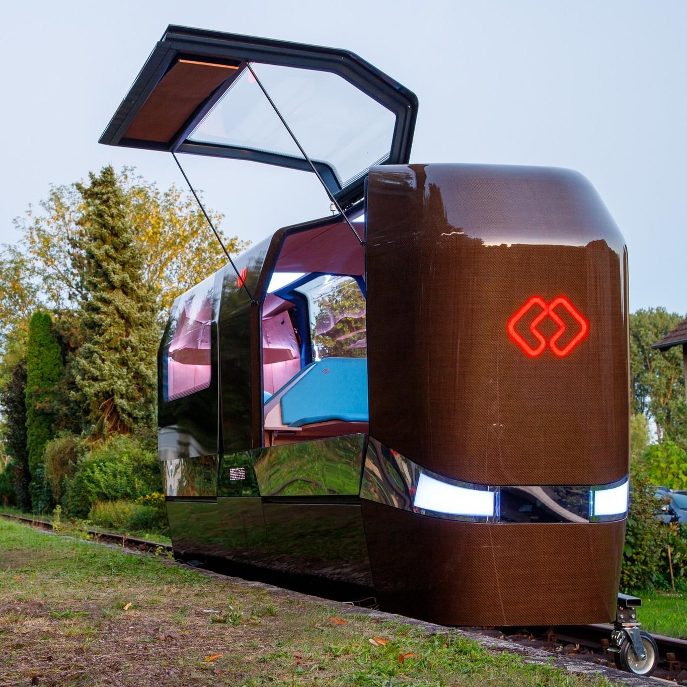 This image shows a gyro-stabilized monorail cabin. The cabin has a reflective, metallic exterior with large windows on the sides with one of them being open and revealing a cozy interior with seating and wood accents. It is situated outdoors on a patch of grass, with residential houses in the background and a clear sky overhead. 