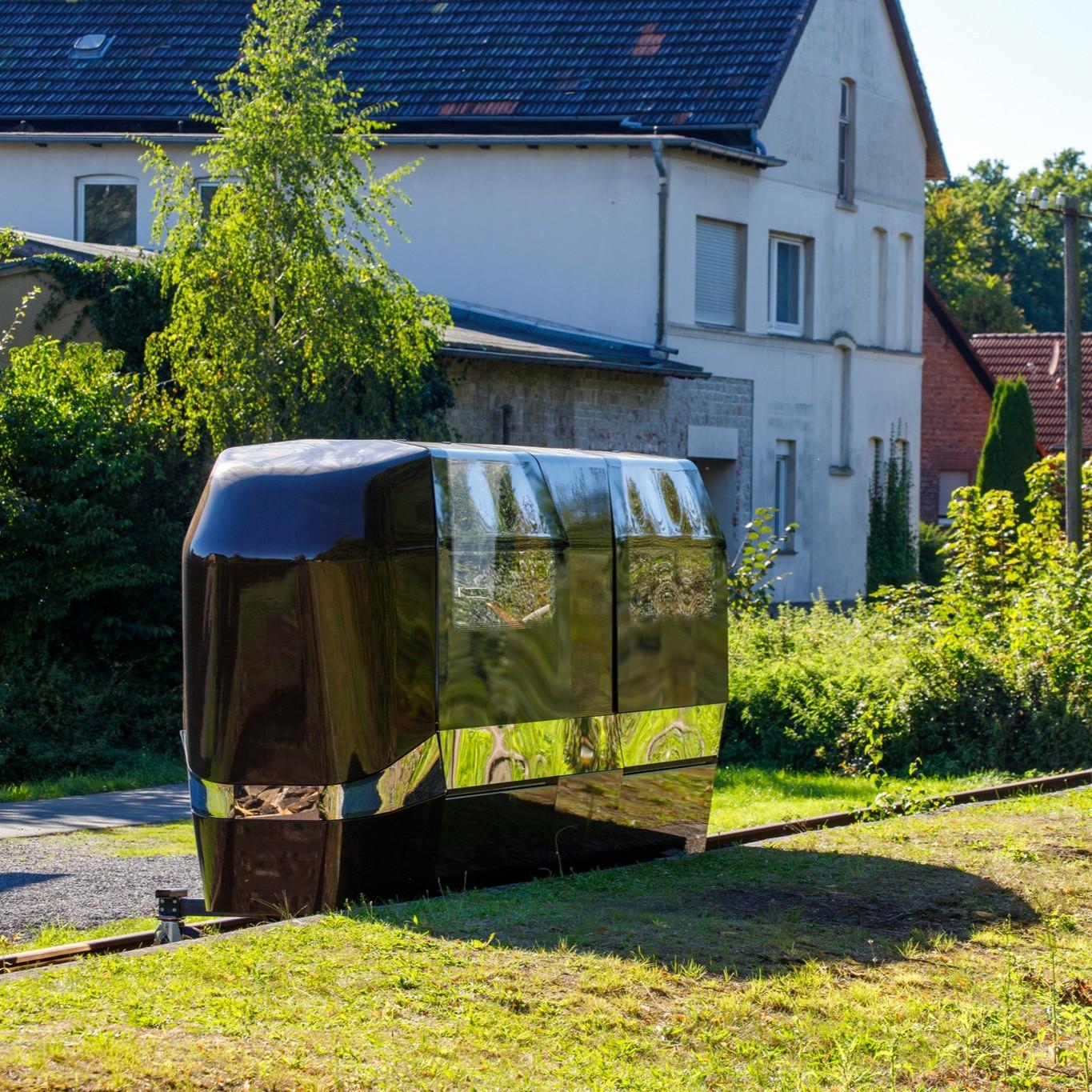 This image shows a sleek, modern, black metallic gyro-stabilized monorail cabin system providing individual, autonomous, and “on-demand” transportation on a small section of railway track in a suburban or rural area. In the background, there are residential houses surrounded by trees and greenery. The scene is brightly lit with sunny weather. 