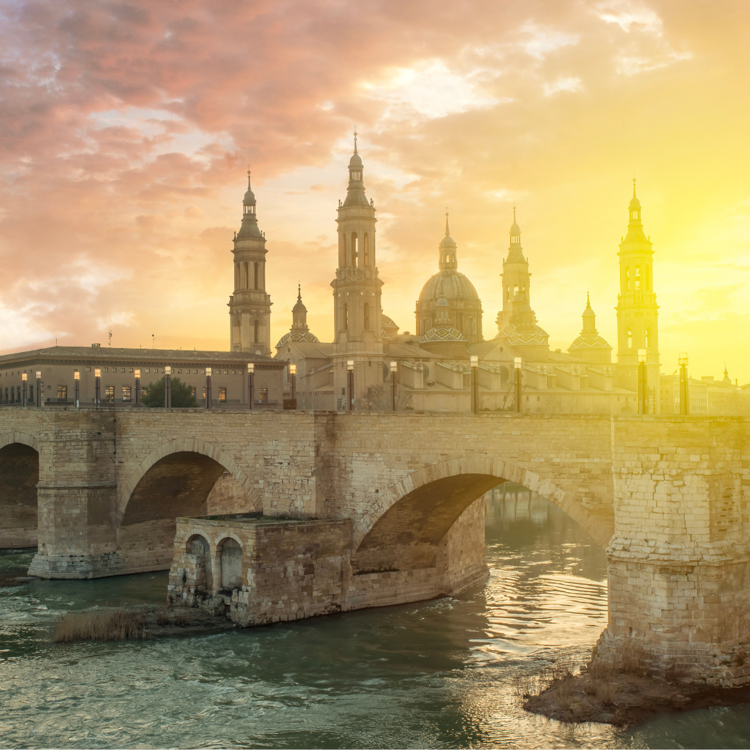 A sunset view of the Puente de Piedra (Stone Bridge) spanning a river in Zaragoza, Spain, with the towering spires and domes of the Basilica de Nuestra Señora del Pilar visible on the far bank under a dramatic orange and yellow sky.