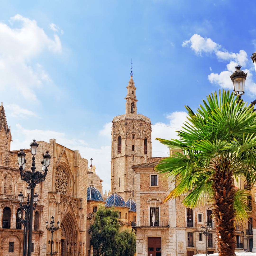 A sunlit photo of the historic Valencia Cathedral complex in Valencia, Spain, featuring a tall bell tower (Miguelete Tower) and an ornate Gothic portal, framed by lampposts and a large palm tree in the foreground.