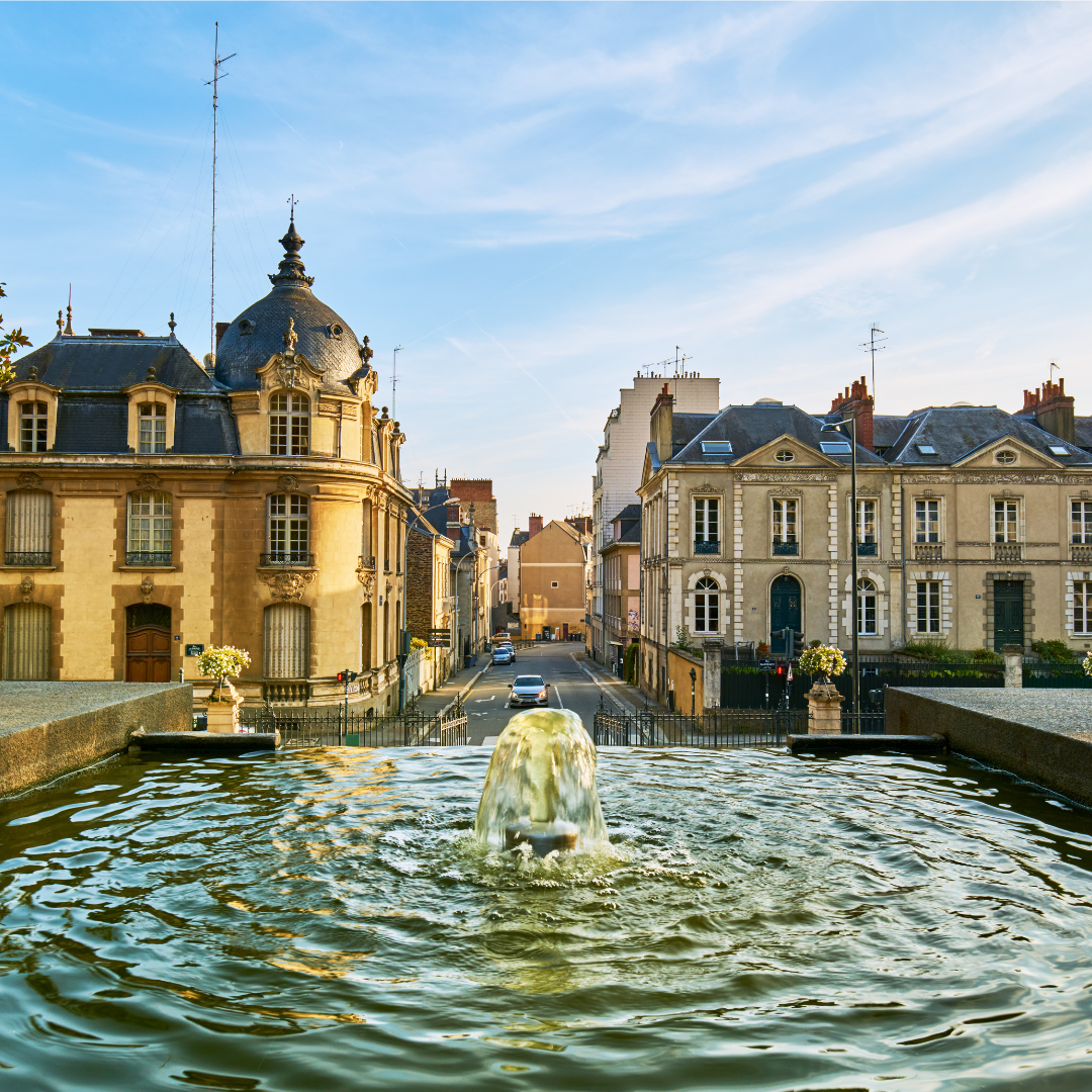 A view over a stone fountain toward a narrow street lined with classic European buildings in Rennes, France. The street leads downhill and away from the viewer.