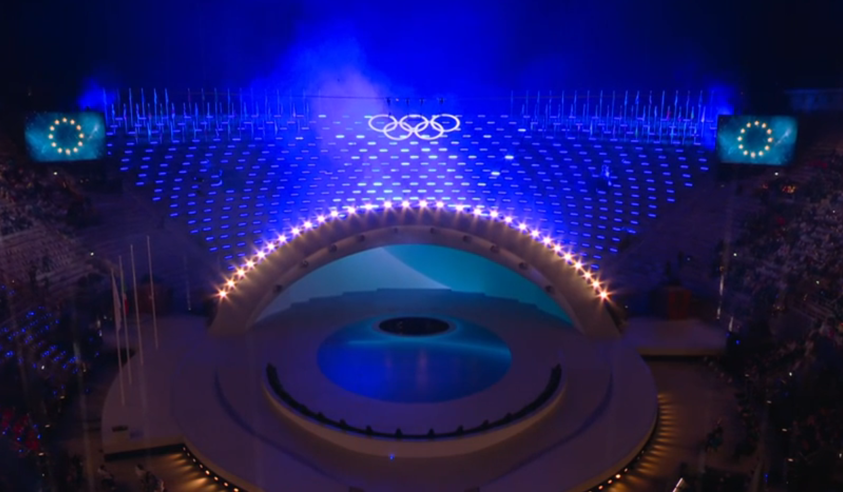 A stadium stage illuminated in blue during an Olympic ceremony, with the Olympic rings above a curved central platform, audience seating on both sides, and European Union flags shown on large screens.
